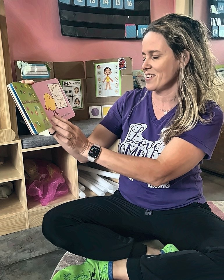 Angela sitting on a classroom rug, smiling as she holds up a picture book