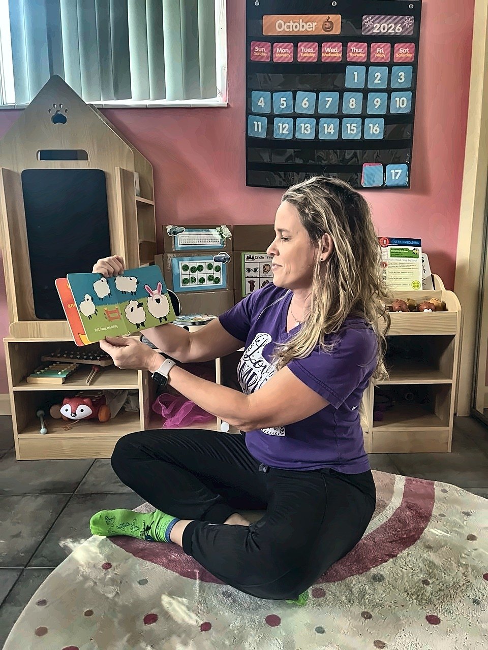 Angela sitting cross-legged on a rug in a classroom, reading a picture book